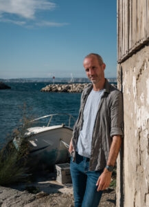 Photo portrait d’un homme au bord de l’eau, calanques de Marseille en fond.